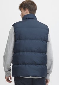 Temno modra napihljena jopica z visokim ovratnikom, kviltanim dizajnom in lahkotno tkanino. Nosil se je nad sivo dolgo srajco z gladko teksturo.