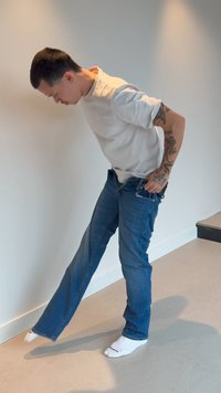 Mężczyzna w białej koszuli i niebieskich dżinsach podciąga dżinsy jedną ręką, balansując na jednej nodze w minimalistycznym wnętrzu.