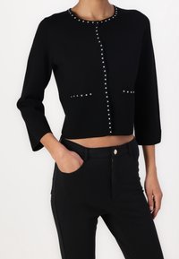 Black cropped cardigan with three-quarter sleeves, featuring silver studs along the neckline and front pockets, made from a smooth, soft material.