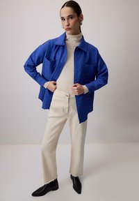 Giovane che indossa una camicia azzurra a bottoni sopra un dolcevita beige, pantaloni crema e stivaletti neri, in piedi contro uno sfondo semplice.