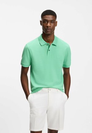 Hombre con polo de manga corta color verde menta y pantalones cortos blancos hasta la rodilla, de pie con las manos en los bolsillos frente a un fondo blanco.