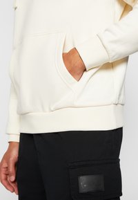 Světle krémová mikina s přední kapsou, vroubkovanými manžetami a spodním lemem. V kombinaci s černými cargo kalhotami se strukturovanou kapsou.