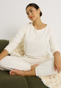 Conjunto de ropa de estar color crema con un patrón floral texturizado. Presenta mangas largas y cintura elástica. Modelo sentado en un suave sofá verde.