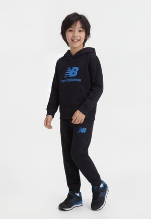 Ragazzo sorridente, indossa una felpa con cappuccio e pantaloni neri New Balance con loghi blu, e sneakers abbinate nere e blu, in piedi su sfondo bianco.