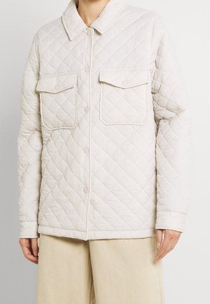 Woman wearing a light beige quilted jacket with front pockets and snap buttons, paired with light beige wide-leg pants.
