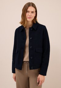 Kvinna som bär en marinblå knappad texturerad jacka över en beige topp och taupe byxor med dragsko, stående mot en beige bakgrund.