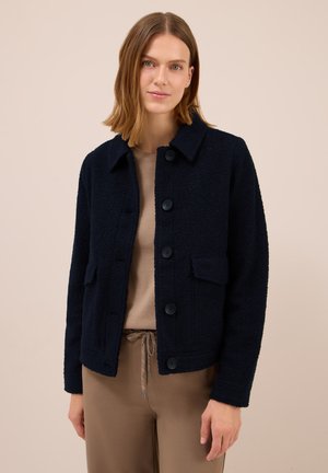 Frau, die eine navyblaue, strukturierte Jacke mit Knöpfen über einem beigen Oberteil und taupefarbene Jogginghose trägt, steht vor einem beigen Hintergrund.