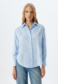 Camisa de botón azul claro con dobladillo redondeado, que presenta diseños florales bordados en tono sobre tono y un cuello clásico. Mangas largas con un ajuste relajado.