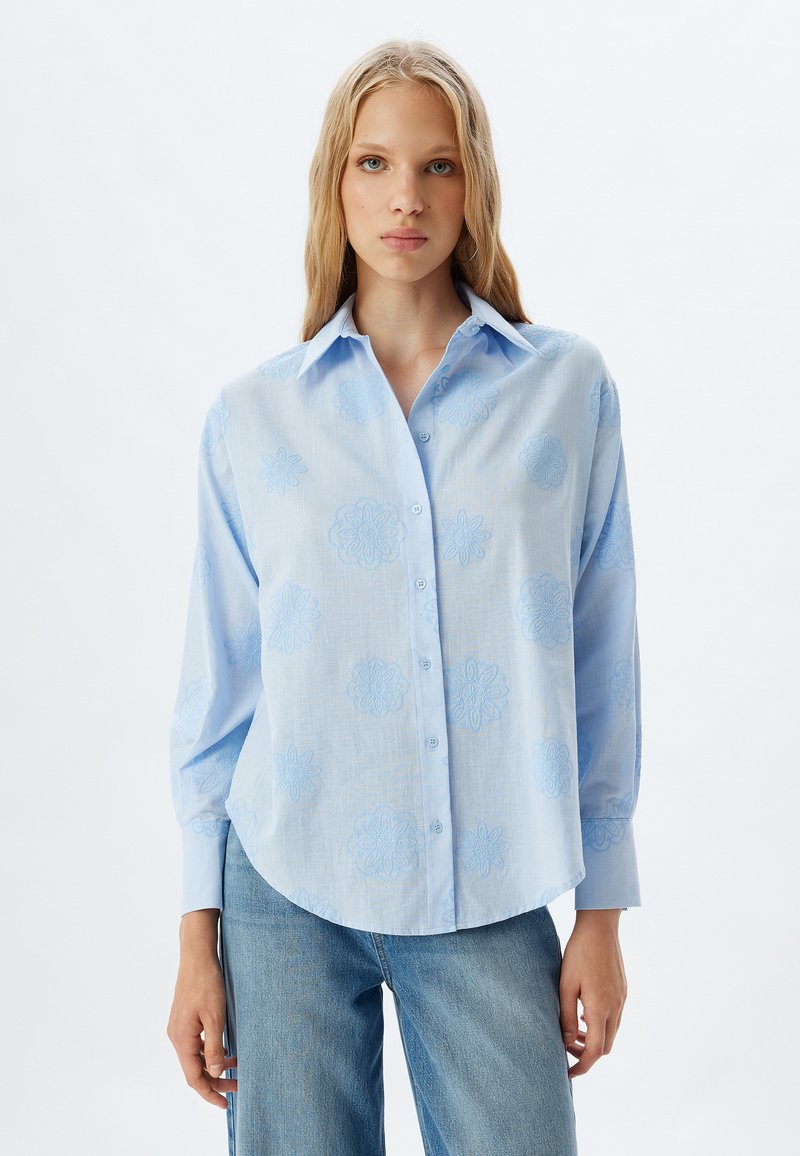 Camicia azzurra chiaro con orlo rotondo, caratterizzata da ricami floreali tonalizzati e colletto classico. Maniche lunghe con vestibilità rilassata.