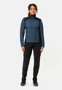 Mėlyna paminkštinta striukė su aukštu apykakliu, juodais peties akcentais, užtrauktuku, tekstūruota paviršiaus, derinama su juodomis kelnėmis ir tamsiais batais.