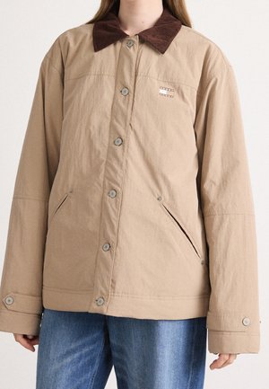 Person wearing a beige buttoned jacket with brown collar and side pockets, paired with blue denim jeans, cropped at shoulders and waist.