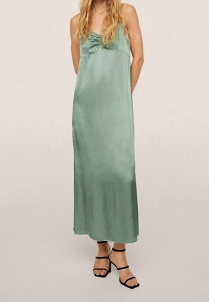 Kobieta ubrana w bezrękawową, sięgającą podłogi sukienkę z zielonego satynu w kolorze szałwii, z marszczeniami na biuście oraz czarne sandały na obcasie z paskami.