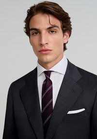 Traje gris oscuro con solapas de muesca; camisa blanca debajo; corbata a rayas burdeos; pañuelo de bolsillo visible en la chaqueta del traje.