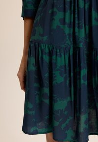 Sukienka w kwiaty w ciemnozielonym i granatowym kolorze. Posiada warstwowy dół oraz krótkie rękawy, wykonana z lekkiego, lekko fakturowanego materiału.