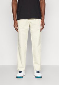 Ανοιχτό μπεζ chinos από βαμβάκι με λεία υφή. Καρφωτό κόψιμο με πλαϊνές τσέπες, συνδυασμένο με λευκά αθλητικά παπούτσια με μπλε λεπτομέρειες.