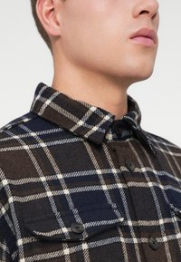Camicia a quadri marrone e blu, realizzata in tessuto flanella, con colletto appuntito, due tasche sul petto e bottoni neri.