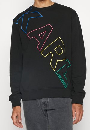 Person trägt einen schwarzen Sweatshirt mit großen mehrfarbigen Umrissbuchstaben, die "KARL" diagonal über die Vorderseite schreiben.