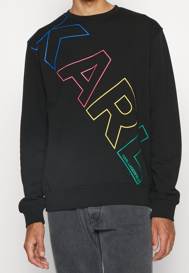Personne portant un sweat-shirt noir avec de grandes lettres multicolores en contour formant le mot "KARL" en diagonale sur le devant.