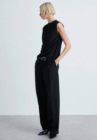 Haut sans manches noir avec un détail drapé, associé à un pantalon noir à jambes larges et des chaussures noires pointues, le tout sur un fond gris clair.