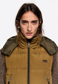 Mujer con cabello oscuro que lleva un chaleco acolchado verde oliva con bolsillos con cremallera y un cuello abotonado sobre un suéter marrón.