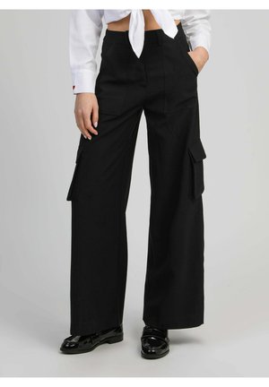 Femme portant un pantalon cargo noir à jambes larges, des mocassins noirs en cuir verni, et un crop top blanc noué, la main dans la poche.