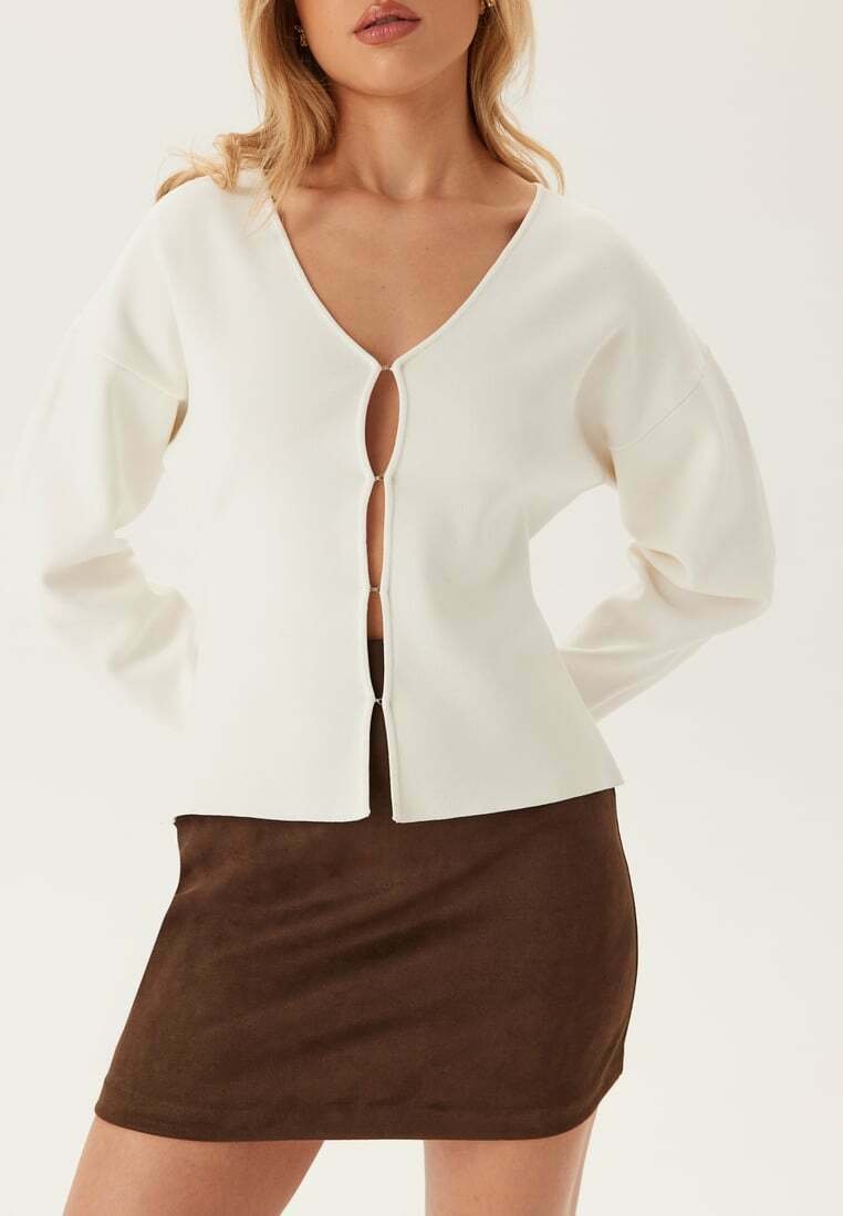 Top bianco lavorato a maglia con scollo a V, dettagli con bottoni sul davanti e maniche corte e morbide, abbinato a una mini gonna in suede marrone. Texture morbida e design casual.