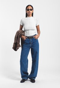 Top blanco ajustado de canalé con cuello alto, combinado con jeans de denim azul de pierna ancha. Suéter marrón colgado del brazo, cinturón negro y gafas de sol.
