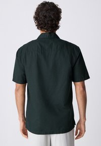 Camisa de manga corta en tela verde oscuro. Presenta un cuello clásico y un corte recto, con una textura suave y sin patrones visibles.