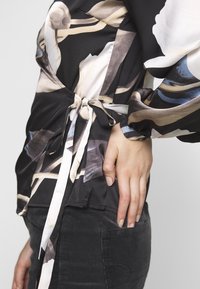 Zwarte blouse met bloemenprint, met lange mouwen en een strikdetail rond de taille. Glad materiaal met een licht glanzende afwerking.