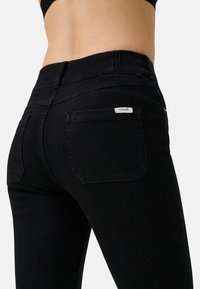 Jeans in denim nero con un design aderente, caratterizzati da una tasca posteriore a toppa e una piccola etichetta con il marchio sul cinturino. Texture liscia.