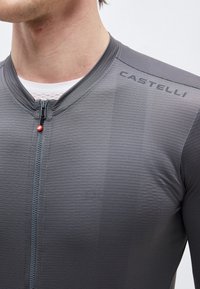 Κοντινό πλάνο ενός ατόμου που φοράει γκρι ποδηλατικό τζέρσεϊ Castelli με μπροστινό φερμουάρ και λευκό διχτυωτό εσώρουχο ορατό στο λαιμό.