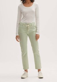 Kvinna iklädd ljusgröna raka byxor med dragsko i midjan, vit långärmad åtsittande topp och vita sneakers, stående mot en enhetlig bakgrund.