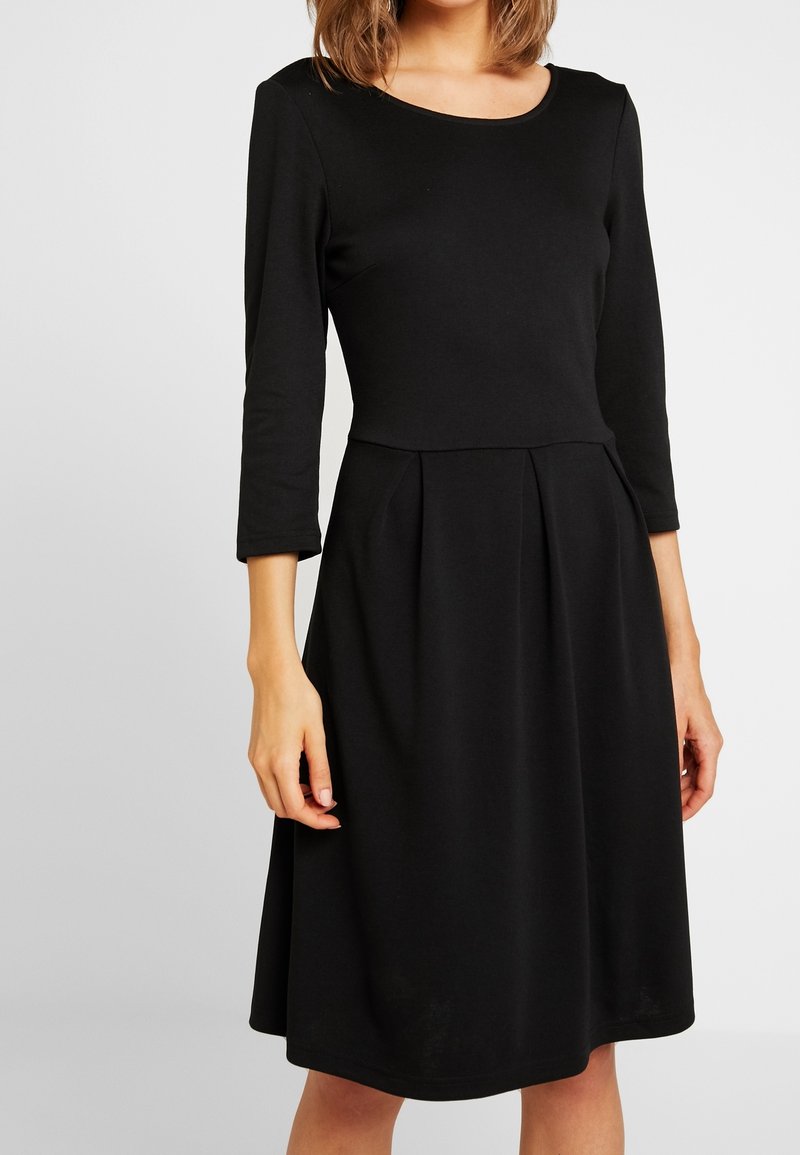Femme portant une robe noire longueur genou avec des manches trois-quarts et un corsage ajusté avec des plis à la taille.