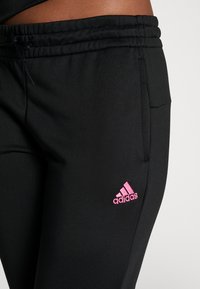 Čierne športové nohavice z hladkej tkaniny s elastickým pásom. Obsahujú ružové logo Adidas na ľavej strane a bočné vrecko.