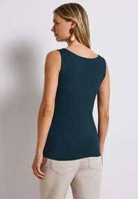 Top senza maniche di colore teal realizzato in morbido tessuto, con scollatura rotonda e forma aderente. Indossato con pantaloni chiari e strutturati.