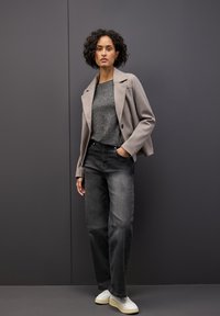 Giacca in misto lana grigio chiaro sopra un maglione grigio scuro, abbinata a jeans in denim neri a vita alta e sneakers bianche. Capelli ricci. Sfondo neutro.