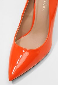 Chaussure à talon haut en cuir verni orange vif, à bout pointu, avec une semelle intérieure beige et une finition brillante sur fond blanc.