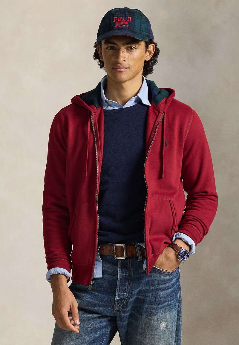 Sweatshirt vermelha com fecho de correr e capuz forrado com padrão xadrez verde, por cima de um suéter azul-marinho e uma camisa azul com colarinho; combinada com jeans escuros e um cinto castanho.