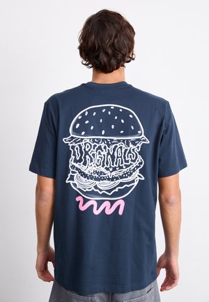 Mand iført navyblå T-shirt med stor hvid kontur af en burger og teksten "Originals" samt en lyserød krusedulle på bagsiden.