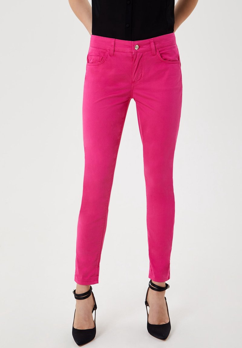 Femme portant un pantalon slim rose vif et des chaussures à talons hauts avec bride à la cheville, posant devant un fond uni.