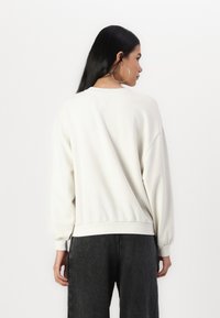 Felpa bianca con scollatura a girocollo, spalle morbide e polsini a costine. Le maniche sono leggermente bombate e presentano una vestibilità ampia.