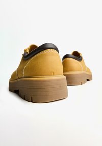 Pantofi din suede galben cu un design rotund, având un accent maro închis în jurul gulerului și o talpă din gumă texturată, cu striații.