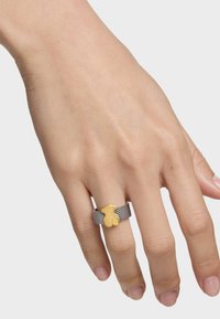 Charm en forma de oso dorado sobre un anillo de malla de plata texturizada, exhibido en una mano con un fondo neutro. Acabado liso y pulido.