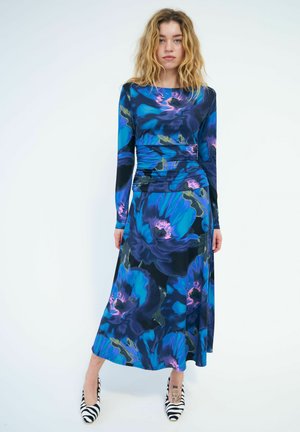 Jeune femme aux cheveux blonds ondulés, portant une longue robe bleue fleurie et des talons hauts à rayures zèbres, debout devant un fond blanc uni.