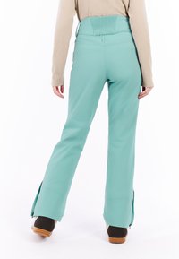 Persoon in turquoise sneeuwbroek en beige lange mouwen top, gezien vanaf de achterkant, staand met de voeten iets uit elkaar op een witte achtergrond.