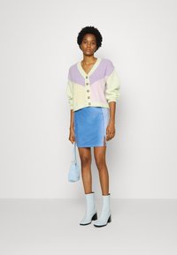 Cardigã de malha com secções em verde pastel, roxo e amarelo, apresentando botões grandes. Saia mini em camurça azul com abertura lateral. Botas de tornozelo em azul claro.