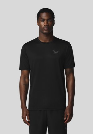 Man staat met het gezicht naar voren, draagt een zwart T-shirt met korte mouwen met een klein vleugellogo op de linkerborst tegen een lichtgrijze achtergrond.