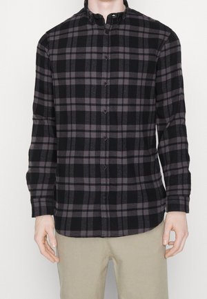 Homme portant une chemise noire et grise à carreaux à manches longues boutonnée, avec un pantalon beige, visible du cou aux hanches, debout devant un fond uni.