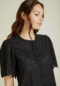 Blouse noire à œillets avec des manches courtes bouffantes, un devant boutonné et de délicats motifs floraux. La texture est légère avec des bords festonnés.