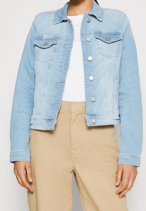 Femme portant une veste en jean bleu clair sur un top blanc et un pantalon beige, debout devant un fond uni.